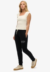 Weißes, enges Oberteil mit gewelltem Saum und quadratischem Ausschnitt, kombiniert mit schwarzen Jogginghosen mit Logoprint und Kordelzug. Beigefarbene Sneakers.