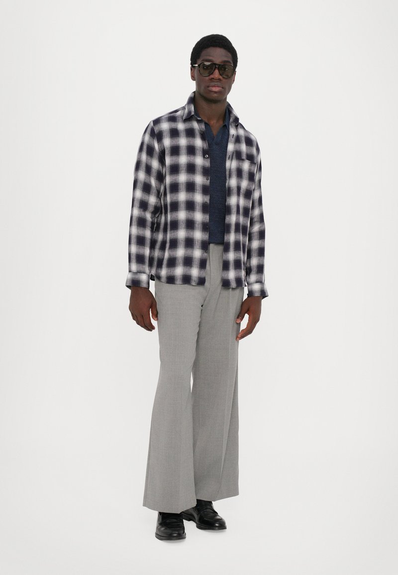 Geruite flanellen shirt in zwart-wit, gelaagd over een marineblauw sweater, gecombineerd met lichtgrijze wijde broek en zwarte schoenen.