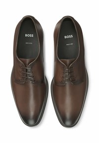 Scarpe in pelle marrone con finitura liscia, punta arrotondata e design con lacci. Presentano un branding discreto all'interno e una suola testurizzata.