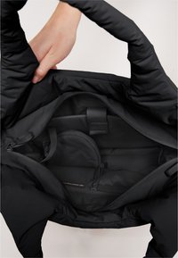 Svart vadderad tote väska med en slät yttre textur, utrustad med ett rymligt innerfack och en liten ficka med dragkedja. Handtag högst upp.