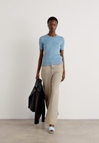 Maglione azzurro chiaro a maglia cable con maniche corte, pantaloni beige a palazzo e sandali blu chiaro. Il modello indossa una giacca nera di pelle.