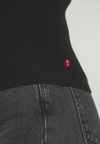 Svart ribbad stickad topp med en röd logotagg, kombinerad med mörkgrå denim. Tyget är texturerat och visar fina vertikala linjer.