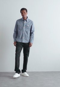 Camisa gris de botones con dos bolsillos en el pecho y botones en tono cobre, combinada con pantalones negros y zapatillas blancas sobre un fondo neutro.