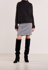 Schwarzer Strickpullover mit lockerer Passform und hohem Kragen. Rock mit schwarz-weißen Muster. Über-die-Knie schwarze Wildlederstiefel mit klobigen Absätzen.