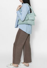 Borsa in pelle verde menta con design annodato. L'utente indossa una camicia azzurra chiara, pantaloni larghi marroni e scarpe slip-on bianche.