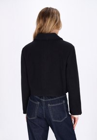 Frau mit langen Haaren, die eine schwarze cropped Jacke und dunkelblaue Jeans trägt, steht mit dem Rücken zu einem weißen Hintergrund.