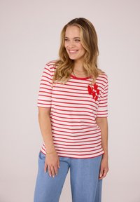 Rot-weiß gestreifes T-Shirt mit kurzen Ärmeln, rundem Halsausschnitt und einem gestrickten roten Herzdetail auf der linken Brust. Kombiniert mit hellblauen Jeans.