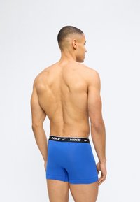 Μυώδης άνδρας με κοντά μαλλιά στέκεται με την πλάτη προς τα εμπρός, φορώντας μπλε εσώρουχα boxer της Nike με μαύρη ζώνη, σε απλό ανοιχτό φόντο.