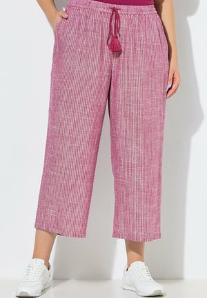 Kvinde iført lyserøde og hvide vertikalt stripede vidde bukser med snøre og hvide sneakers, stående med hænderne i lommerne.