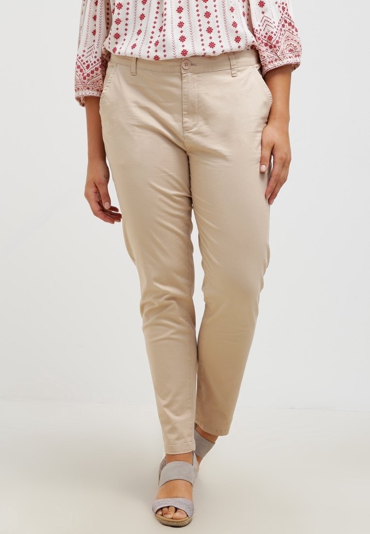 Femme portant un pantalon beige slim, associé à un chemisier blanc et rouge à motifs et des sandales grises à bouts ouverts, debout devant un fond uni.