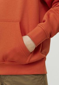 Felpa arancione con cappuccio, dotata di tasca frontale e polsini a coste. Texture liscia e design casual. Indossata sopra pantaloni khaki.