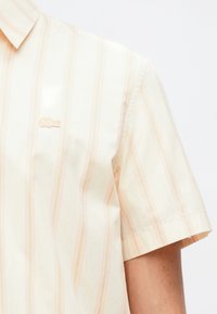 Κοντομάνικο ανοιχτό κίτρινο πουκάμισο με ανοιχτό ροζ κατακόρυφες ρίγες, που διαθέτει ένα μικρό κεντημένο λογότυπο στο στήθος. Λείος βαμβακερός υφασμάτος.