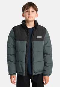 Ragazzo che indossa un giubbotto imbottito nero e grigio con zip frontale e una maglietta blu navy sotto, in piedi su uno sfondo bianco.