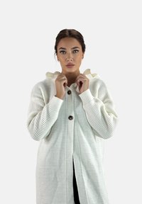 Cárdigan blanco de punto con capucha, que presenta un patrón texturizado, puños acanalados y dos grandes botones en la parte delantera.