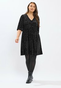 Schwarzes Samtkleid mit sternförmigen silbernen Sprenkeln, V-Ausschnitt, ärmellange Ärmel und gefaltete Taille. Getragen mit schwarzen Strumpfhosen und Knöchelstiefeln.