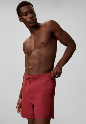 Hombre sin camisa que lleva pantalones cortos de baño rojos, tirando de la cintura, mirando hacia abajo, de pie frente a un fondo gris liso.