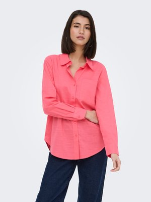 Femme portant une chemise rose vif à manches longues boutonnée et un pantalon bleu foncé, debout avec un bras croisé sur la taille.
