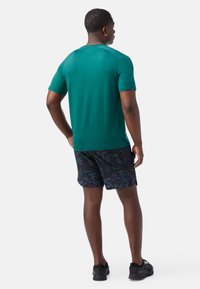 Maglietta atletica turchese a maniche corte, abbinata a pantaloni corti neri con un motivo astratto multicolore. Il modello è in piedi con le spalle rivolte verso la macchina fotografica.