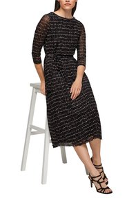 Vestido negro de longitud midi con un patrón de cuadrícula, mangas transparentes y cintura ceñida con cinturón, combinado con tacones negros con tiras. Brillo sutil en la tela.