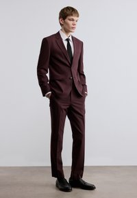 Σκουρόχρωμο μπορντό κοστούμι με ραμμένη σχεδίαση, κούμπωμα με ένα κουμπί, πέτα με εγκοπές, σε συνδυασμό με λευκό πουκάμισο και μαύρη γραβάτα, ολοκληρωμένο με μαύρα μοκασίνια.