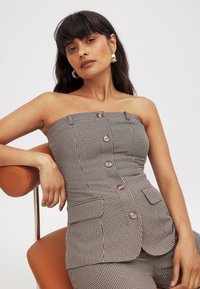 Top bez ramienok s károvaným vzorom v hnedej a smotanovej farbe, s piatimi gombíkmi, bočnými vreckami a priliehavým strihom. Modelka odpočíva na oranžovej stoličke.