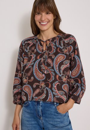 Lächelnde Frau mit braunen Haaren trägt eine braune Bluse mit Paisley-Muster und Schluppe am Hals sowie blaue Jeans und posiert vor einem einfarbigen Hintergrund.