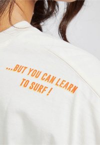 Camiseta de algodón color crema con cuello redondo; presenta un texto en un vibrante color naranja que dice "...PERO PUEDES APRENDER A HACER SURF!" en el hombro de la parte de atrás.