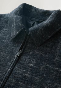 Dunkelgrau Wollsakko mit strukturiertem Stoff, hohem Kragen und teilweise geschlossener sichtbarer schwarzer Reißverschluss.