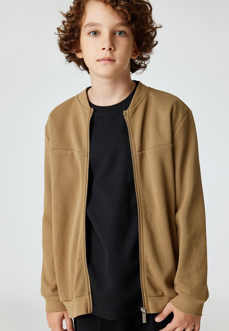 Beige zip-up jack met een gestructureerde afwerking, ronde halslijn en geribbelde manchetten, gelaagd over een zwart shirt met lange mouwen.