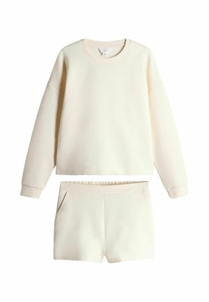 Kreminės spalvos ilgomis rankovėmis megztinis su atitinkančiais šortais, turi ribotą apvalią iškirptę ir elastinę juosmens juostą šortuose.