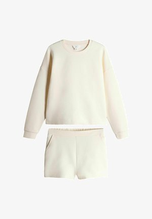 Kreminės spalvos ilgomis rankovėmis megztinis su atitinkančiais šortais, turi ribotą apvalią iškirptę ir elastinę juosmens juostą šortuose.