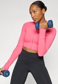 Fitted, cropped long-sleeve top in bright pink, paired with black high-waisted leggings. The person holds a blue dumbbell with a number 3.