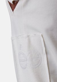 Detailaufnahme einer weißen Kleidungsstücktasche mit strukturiertem Stoff und subtiler gestickter Gestaltung auf der Vorderseite, getragen von einer Person mit der Hand darin.