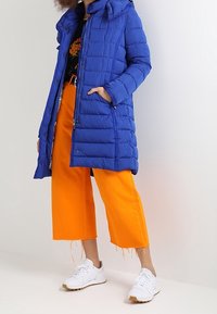 Mujer vistiendo un abrigo acolchado azul, una blusa negra con estampado floral, pantalones cortos de pierna ancha en naranja brillante y zapatillas blancas, de pie con una mano en el bolsillo.
