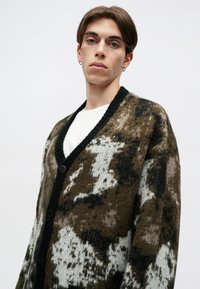 Pleteni kardigan u smešnoj, crnoj i beloj kamuflažnoj šemi, sa crnim V-izrezom, dugmadi za zatvaranje i mekanom teksturom.