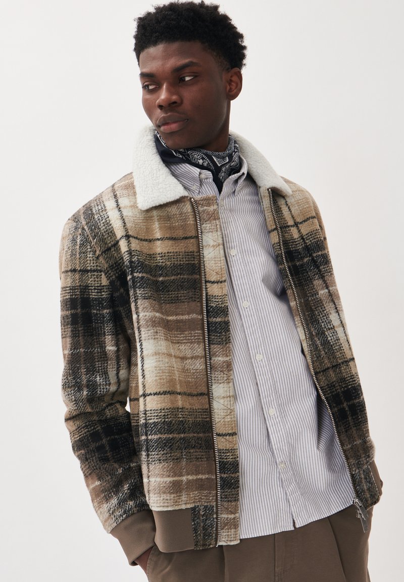 Karierte Bomberjacke in Braun- und Beigetönen mit einem Kunstfellkragen. Verfügt über einen Frontreißverschluss und gerippte Manschetten. Getragen über einem gestreiften Hemd.