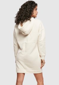 Kapuci džemperkleita gaiši bēšā krāsā. Tai ir garas piedurknes, mīksta tekstūra un brīva dizaina elements ar šuvi aizmugurē.