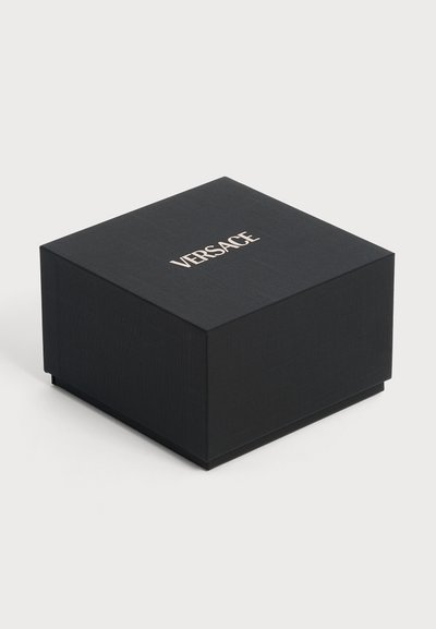 Boîte carrée noire Versace avec surface texturée et nom de la marque blanc centré sur le couvercle, présentée sur un fond clair uni.