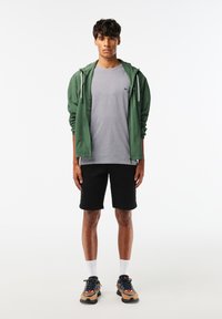 Grön lättvikts zip-jacka, grå t-shirt, svarta shorts och flerfärgade sneakers med vita strumpor; modell som står mot en neutral bakgrund.