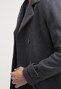 Hombre con un abrigo de lana gris oscuro con botones negros, sujetándose el abrigo cerrado a la altura del pecho sobre un fondo claro y liso.