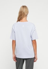 Blond persoon die met zijn rug naar de camera staat, gekleed in een lichtpaars oversized T-shirt met een klein wit Jumpman-logo op de bovenrug, gecombineerd met grijze broek.