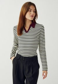 Melnā un baltā svītrainā ilga piedurkņu krekls ar bordo V veida apkakli, kombinēts ar augsto vidukli melniem biksēm ar divu pogu dizainu.