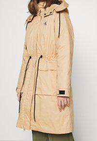 Light beige parka jacket with an adjustable drawstring waist, hood, front pockets, logo on the chest, and black accent details.