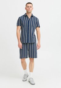 Kortærmet skjorte og shorts sæt i mørkeblå med lodrette hvide og creme striber. Fremstillet af et tekstureret, åndbart stof. Har en lomme.