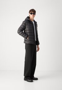 Giacca a vento nera con cappuccio e design trapuntato. Abbinata a pantaloni neri e scarpe nere, il modello indossa occhiali da sole. Maglia grigio chiaro sotto.