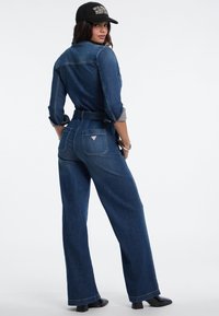 Donna che indossa una tuta di denim blu scuro, scarpe con tacco nere e un cappellino nero girato all'indietro su uno sfondo bianco semplice.