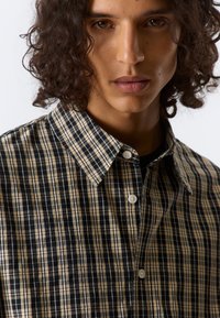 Chemise à carreaux noire, blanche et beige. Dotée d'un col évasé, de manches courtes et de boutons blancs. La texture semble lisse.