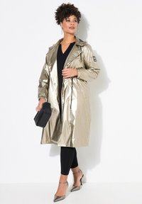 Metalen gouden trenchcoat met een koord in de taille, zwarte accenten en een glanzende afwerking. Gecombineerd met zwarte leggings en zilveren hakken.