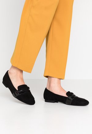 Mocassins en daim noirs avec une bande noire brillante et des détails en frange. Design profil bas, semelle plate, associés à un pantalon jaune.