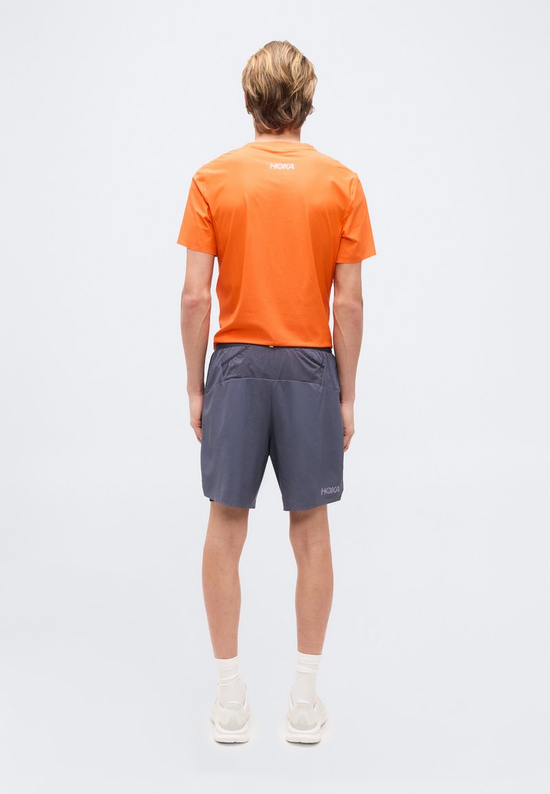 Person, der mit dem Rücken zur Kamera steht, trägt ein oranges Kurzarmshirt, dunkelgraue Shorts, weiße Socken und weiße Turnschuhe vor einem einfarbigen Hintergrund.